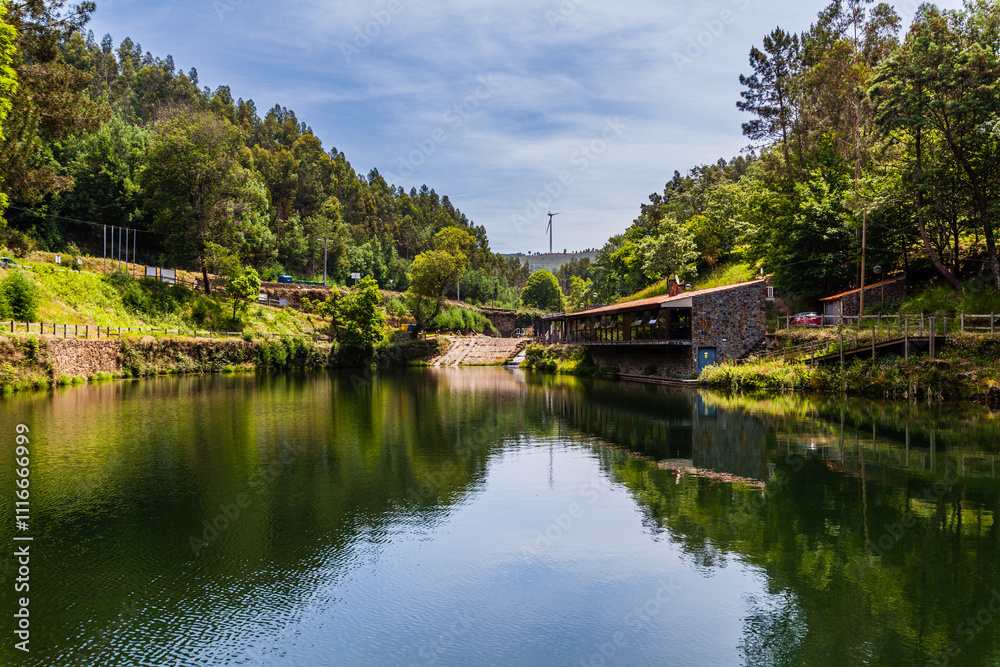 Obraz premium A tranquil lake surrounded by lush green forests, with a small building on the shore and a bridge crossing the water