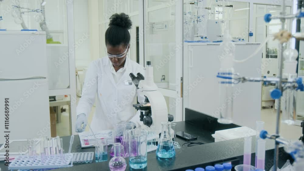 Zoom out shot of African American young female biologist using microscope and writing report for science experiment in modern laboratory