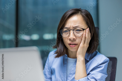 Asian businesswoman wearing glasses looks concerned while working on her laptop in an office setting. Her serious expression conveys focus and determination. Ideal for business-related content.