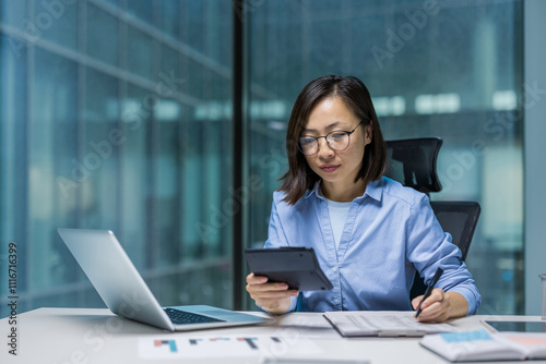 An Asian woman is deeply engaged in accounting work at a modern office desk. She is using a tablet and laptop to manage financial tasks efficiently. Her serious expression reflects concentration