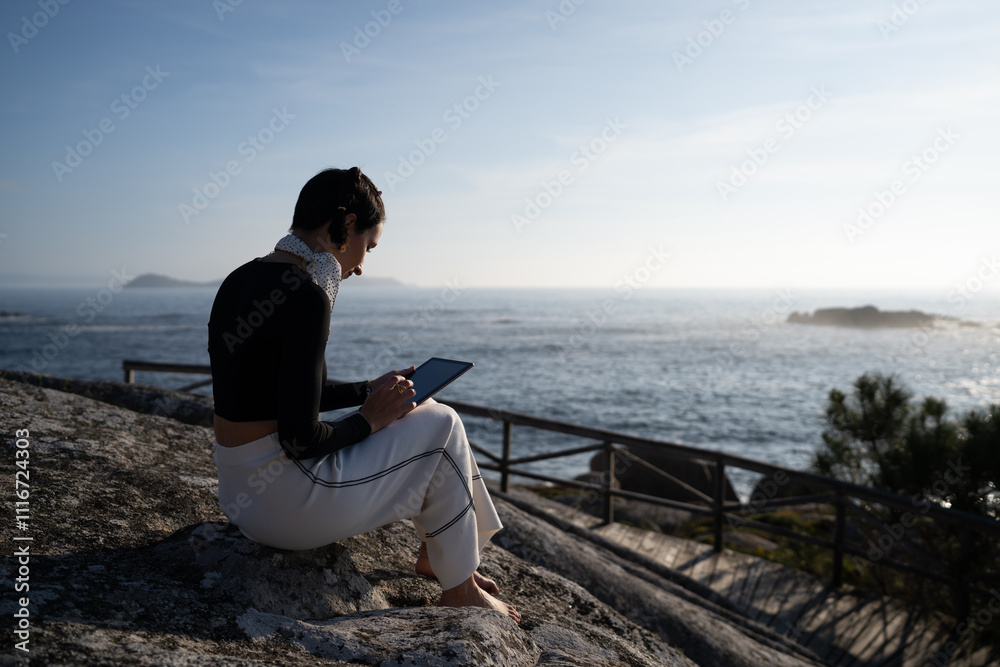 Young woman with bare feet relaxes with her tablet, watching the sunset on a rock by the ocean, creating a peaceful image of work life balance Young woman with bare feet relaxes with her tablet, watching the sunset on a rock by the ocean, creating a peaceful image of work life balance