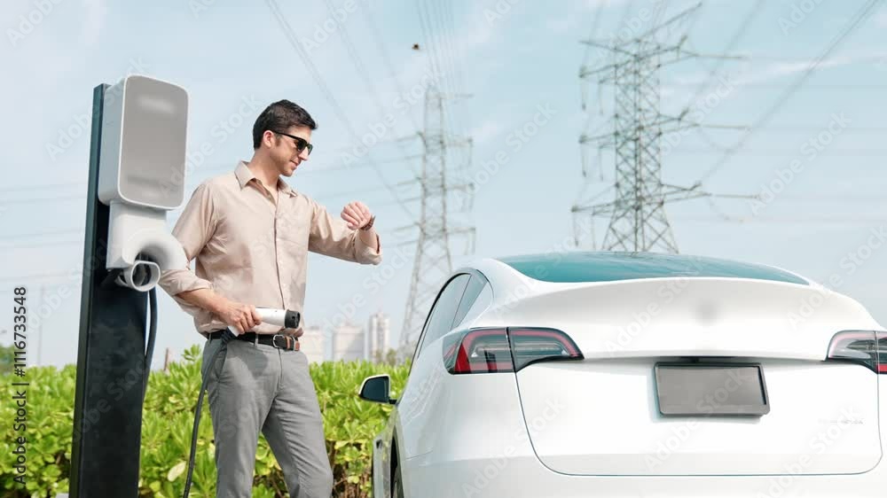 Slow motion young man recharge EV car battery at charging station connected to power grid tower electrical industrial facility as electrical industry for eco friendly vehicle utilization. Expedient Slow motion young man recharge EV car battery at charging station connected to power grid tower electrical industrial facility as electrical industry for eco friendly vehicle utilization. Expedient