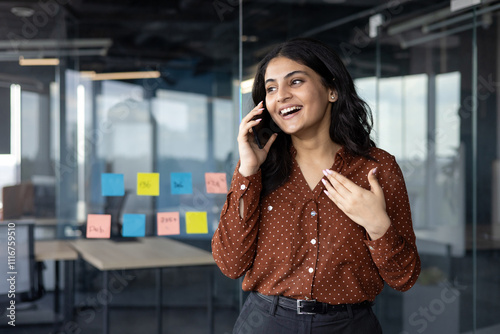 A cheerful businesswoman is engaged in a lively phone conversation in a contemporary office setting, featuring brainstorming notes on glass walls. Her expression reflects positivity