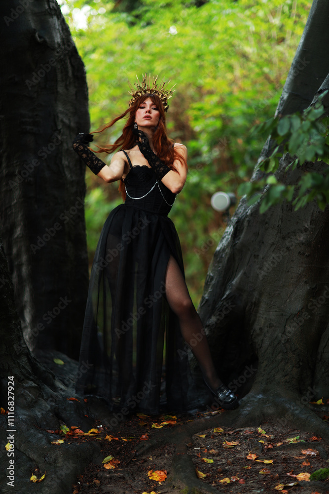 Naklejka premium Full length portrait of young brunette woman in vintage black dress and crown at old trees