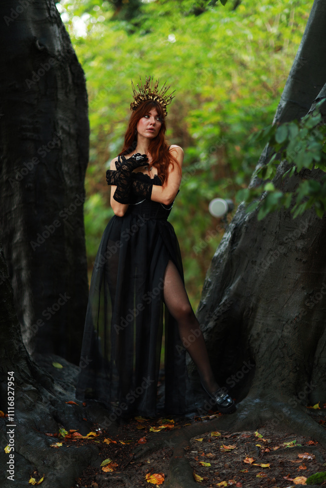 Naklejka premium Full length portrait of young brunette woman in vintage black dress and crown at old trees