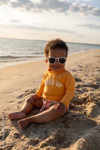 A baby laughing at the camera sitting on the sand at the beach photo