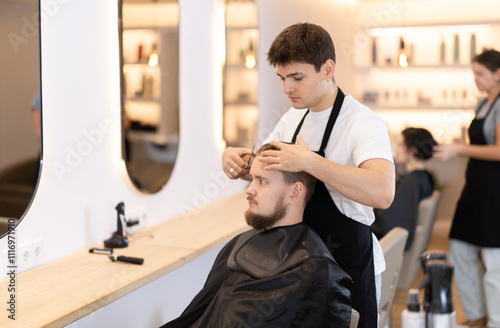 Young man barbershop employee stands next to male client, discussing details of haircut. Stylist and client agree on haircut, master attentively listens to wishes of visitor. photo