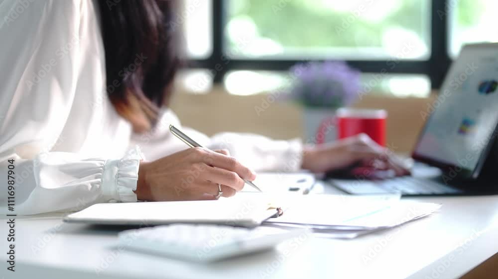 A woman is writing on a piece of paper with a pen. She is sitting at a desk with a laptop and a cup of coffee. The scene suggests that she is working or studying, possibly on a project or assignment