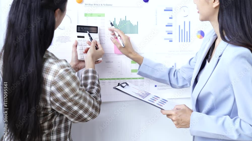 Two businesswomen in a collaborative office environment, analyzing financial data displayed on a whiteboard. They are using a calculator and taking notes