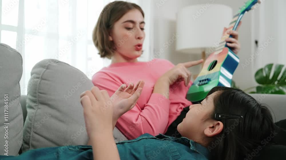 Asian cute daughter listening acoustic music while lie on her mother lap at sofa. Caucasian mother playing colorful ukulele while cute child looking at her parent. Family recreation concept. Pedagogy.
