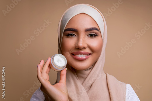 A woman is holding a jar of cream. She is smiling. The cream is white. The woman is wearing a scarf