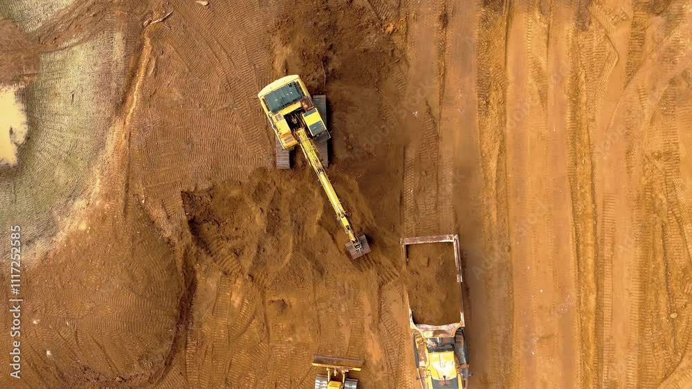 Aerial top down static of excavator clearing dirt as dump trucks approach at construction site, focused on earthmoving machinery