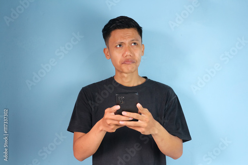 Young Asian man thinking about something while holding his mobile phone