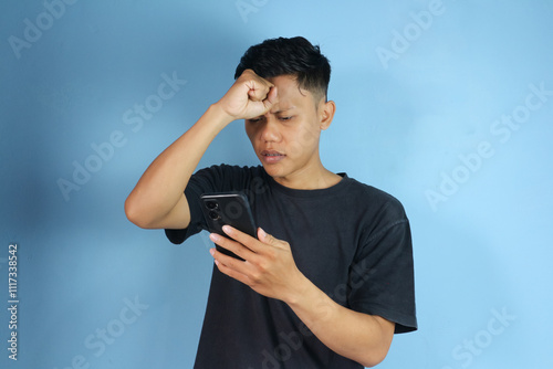 Young Asian man looking at mobile phone with sad expression while holding head 