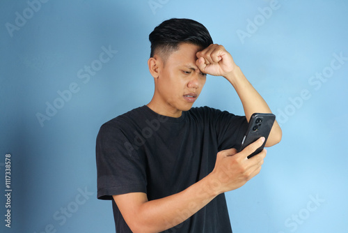 Young Asian man looking at mobile phone with sad expression while holding head 