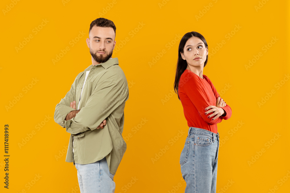 Fototapeta premium A young married couple stands back to back, both with crossed arms, showcasing contrasting emotions. The vibrant yellow background highlights their connection and tension in a playful way.