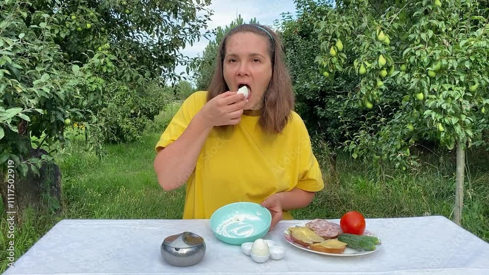 A beautiful young woman enjoys a casual lunch outdoors in a lush green park surrounded by trees. Relaxing and savoring her meal, she takes a moment to unwind in the fresh air and nature.