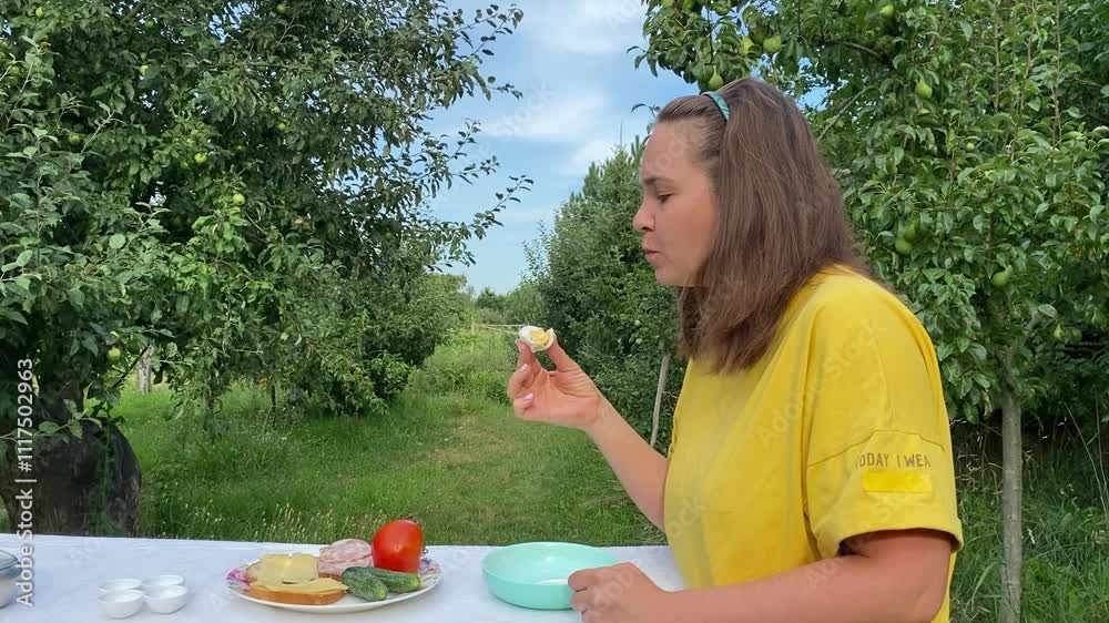 A beautiful young woman enjoys a casual lunch outdoors in a lush green park surrounded by trees. Relaxing and savoring her meal, she takes a moment to unwind in the fresh air and nature.