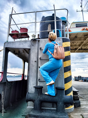 A person in blue climbing a metal ladder on a docked vessel during an overcast day by the waterfront