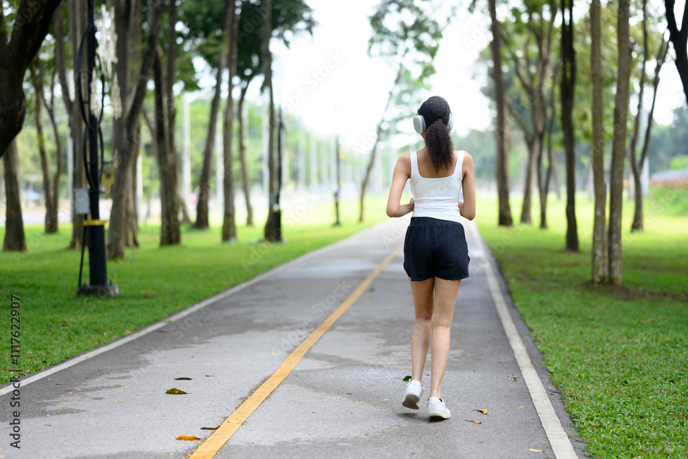 Back view of young woman wearing headphones jogging on pathway in the park Back view of young woman wearing headphones jogging on pathway in the park