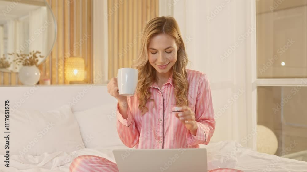 Caucasian woman wearing pink pajama sitting in lotus position on soft bed. Pretty woman using laptop and drinking cup of coffee after sleep. Cheerfully smiling and having good feeling at morning Caucasian woman wearing pink pajama sitting in lotus position on soft bed. Pretty woman using laptop and drinking cup of coffee after sleep. Cheerfully smiling and having good feeling at morning