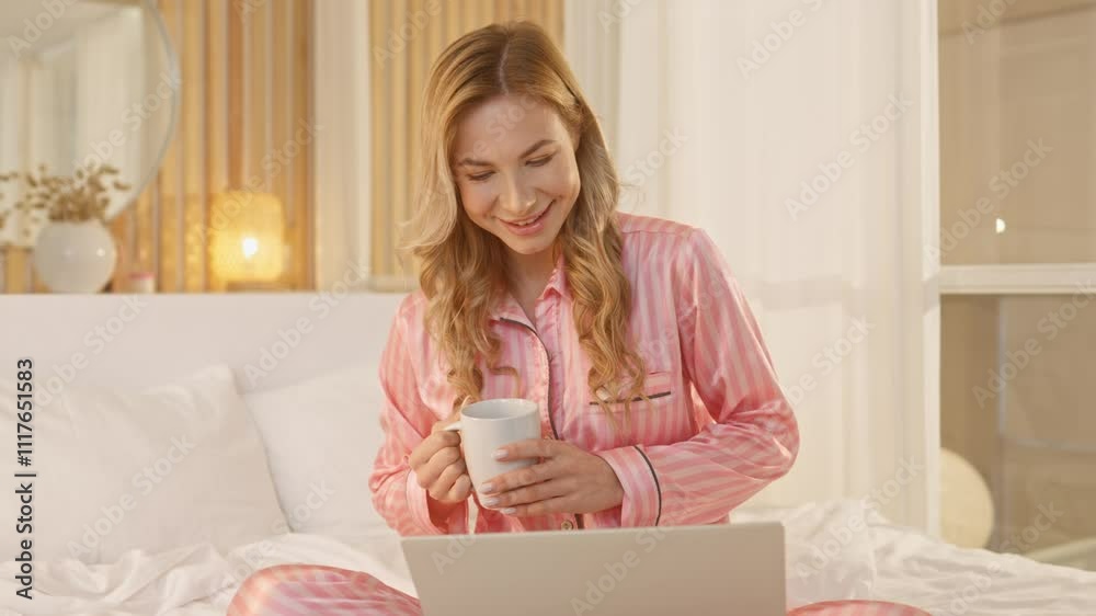Cute Caucasian woman relaxing in lotus position on soft bed. Positive female with stylish pajama drinking cup of coffe or tea. Having good feeling while using laptop. Resting at home. Cute Caucasian woman relaxing in lotus position on soft bed. Positive female with stylish pajama drinking cup of coffe or tea. Having good feeling while using laptop. Resting at home.