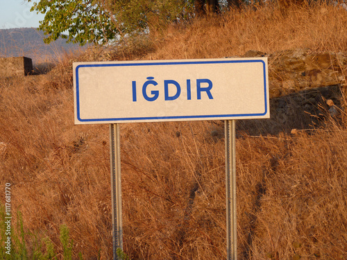 Blue 'Igdir' Sign on a Metal Pole: A Bold Contrast Against the Natural Landscape of Brown Grass and Soft Shadows, Capturing a Serene Afternoon in the Heart of the City.