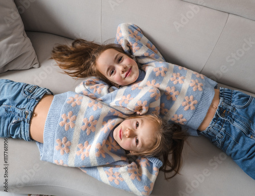 Two adorable sisters, wearing matching pastel sweaters and jeans, playfully lie on a comfy couch. Their smiles and relaxed poses capture the joy of childhood and sibling bonding photo