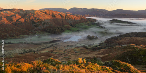 Golden light at sunrise hitting mountain peaks surrounding Little Langdale Tarn as autumn mist fills in the valleys. Lake District, UK. photo