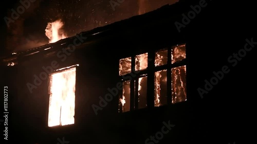 House is on fire at night, flames inside at home, burning windows. The wooden building is ablaze, completely engulfed in fire. Loss of housing, disaster, trouble for unhappy people.