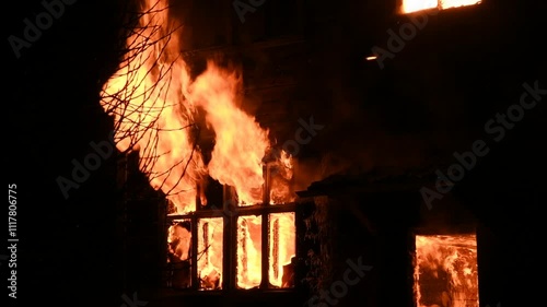 House is on fire at night, flames inside at home, burning windows. The wooden building is ablaze, completely engulfed in fire. Loss of housing, disaster, trouble for unhappy people.