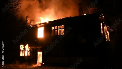 House is on fire at night, flames inside at home, burning windows. The wooden building is ablaze, completely engulfed in fire. Loss of housing, disaster, trouble for unhappy people.