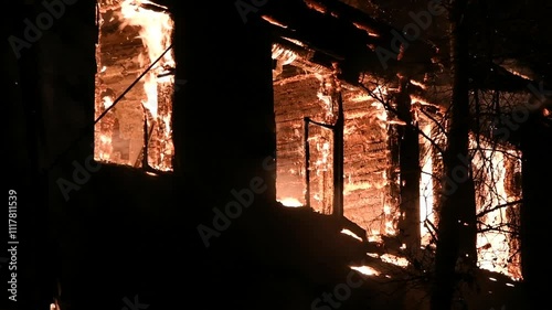 House is on fire at night, flames inside at home, burning windows. The wooden building is ablaze, completely engulfed in fire. Loss of housing, disaster, trouble for unhappy people.