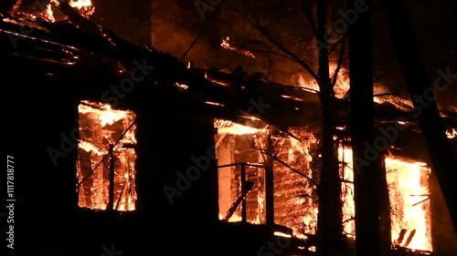 House is on fire at night, flames inside at home, burning windows. The wooden building is ablaze, completely engulfed in fire. Loss of housing, disaster, trouble for unhappy people.