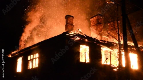 House is on fire at night, flames inside at home, burning windows. The wooden building is ablaze, completely engulfed in fire. Loss of housing, disaster, trouble for unhappy people.