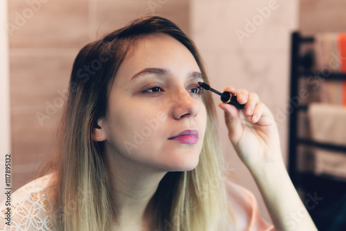 A woman applies mascara to her eyelashes in front of a mirror