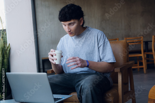 Young man holding his coffee while attentively watching the laptop monitor sitting in the coffee shop