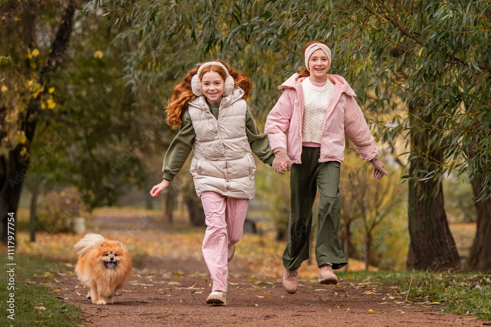 Two happy sister girls run along the path of an autumn park with their pet dog Pomeranian Spitz Two happy sister girls run along the path of an autumn park with their pet dog Pomeranian Spitz