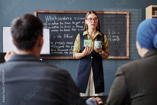 Medium full shot of female English language instructor showing class coffee mug and sticky note with foreign word written while introducing new vocabulary to adult students in classroom, copy space photo