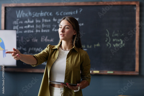 Medium shot of female English teacher holding tablet computer addressing student with question, while discussing new grammar rules in group class against blackboard at language school photo