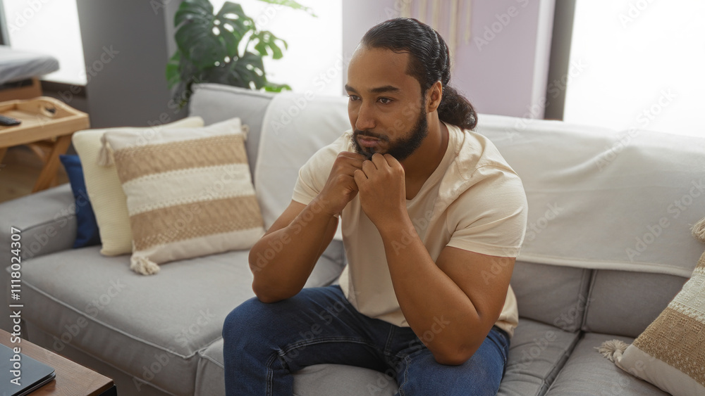 Young man with beard sitting thoughtfully in modern living room interior with cushions and plants, embodying a calm and relaxed atmosphere at home. Young man with beard sitting thoughtfully in modern living room interior with cushions and plants, embodying a calm and relaxed atmosphere at home.