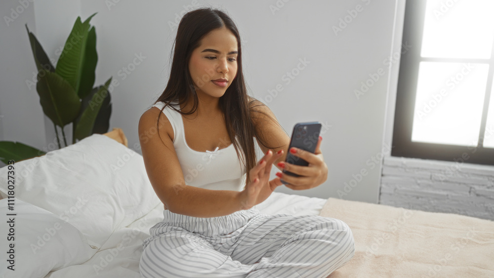 Woman using smartphone while sitting on bed in modern bedroom captures relaxed indoor atmosphere, highlighting her latin beauty with a serene and confident expression. Woman using smartphone while sitting on bed in modern bedroom captures relaxed indoor atmosphere, highlighting her latin beauty with a serene and confident expression.