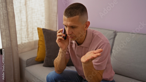 Young man in a living room talking on the phone with a puzzled expression sitting on a sofa with colorful pillows.