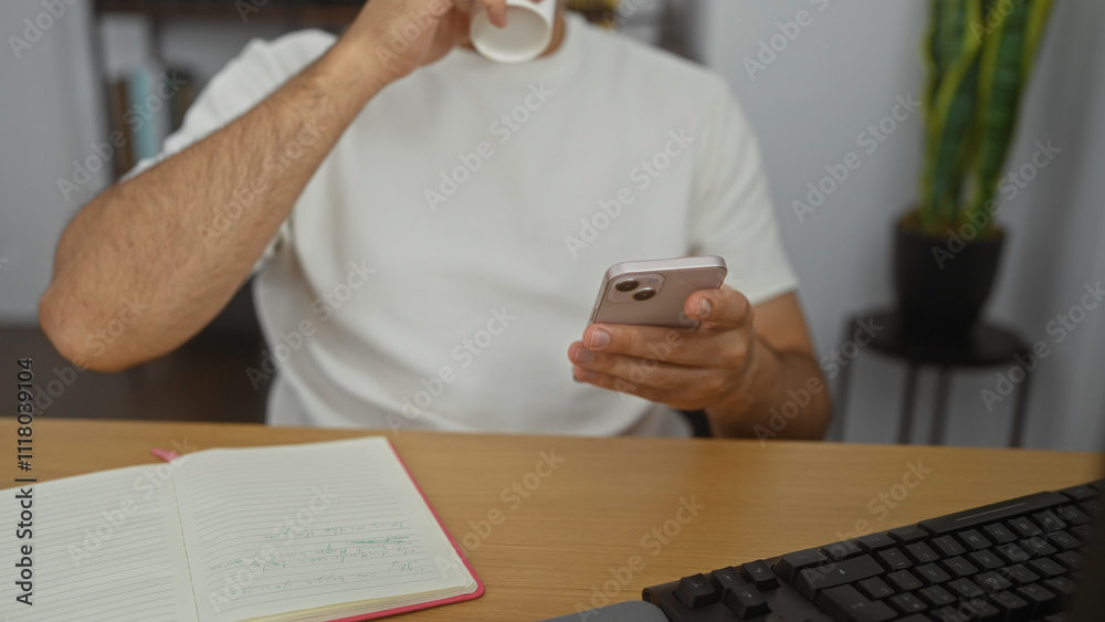 Fototapeta premium Hispanic man in an office sipping coffee while using a smartphone, with hands visible near a notebook on the desk.