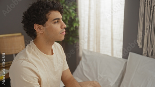 Handsome young hispanic man in a bedroom, sitting on a bed with a relaxed expression, surrounded by soft lighting and cozy decor, creating a serene and comfortable home interior.