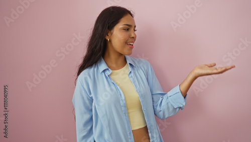 Young woman with an extended hand presenting something, isolated over a pink background, showcasing attractive and beautiful expressions, wearing a blue shirt and a yellow top.