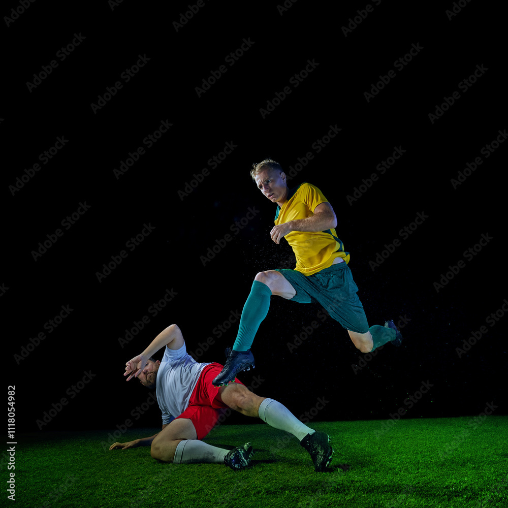 Player in yellow and green uniform jumps over fallen opponent on green football field against black studio background. Concept of sport event, championship, motion, match, game, energy. Ad Player in yellow and green uniform jumps over fallen opponent on green football field against black studio background. Concept of sport event, championship, motion, match, game, energy. Ad
