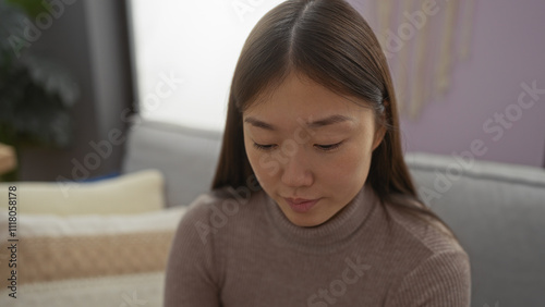 Asian woman sitting in a cozy living room, engaging with a calm demeanor, surrounded by soft furniture, reflecting a peaceful home environment.