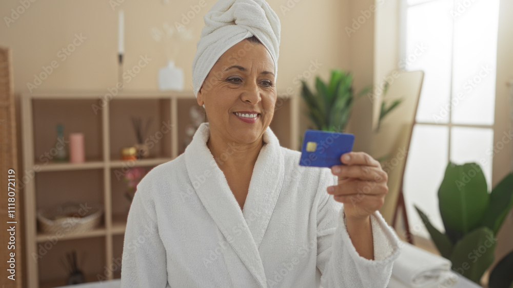 An elderly hispanic woman in a white bathrobe and towel smiling while holding a blue credit card in a serene indoor spa setting surrounded by plants and shelves with relaxation items. An elderly hispanic woman in a white bathrobe and towel smiling while holding a blue credit card in a serene indoor spa setting surrounded by plants and shelves with relaxation items.