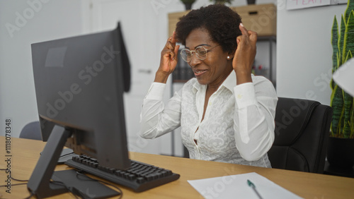 African american woman with curly hair crossing fingers in office setting while sitting in front of computer monitor and desk, wearing glasses and white shirt.